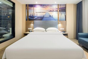 Quarto de hotel com uma cama grande, duas mesas de cabeceira, um criado-mudo, um sofá azul e uma janela ampla.