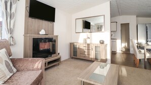A modern living room with a flat-screen TV, a fireplace, a dining area with a table and chairs, and a kitchen visible through an open door. - Draco (Morpeth)
