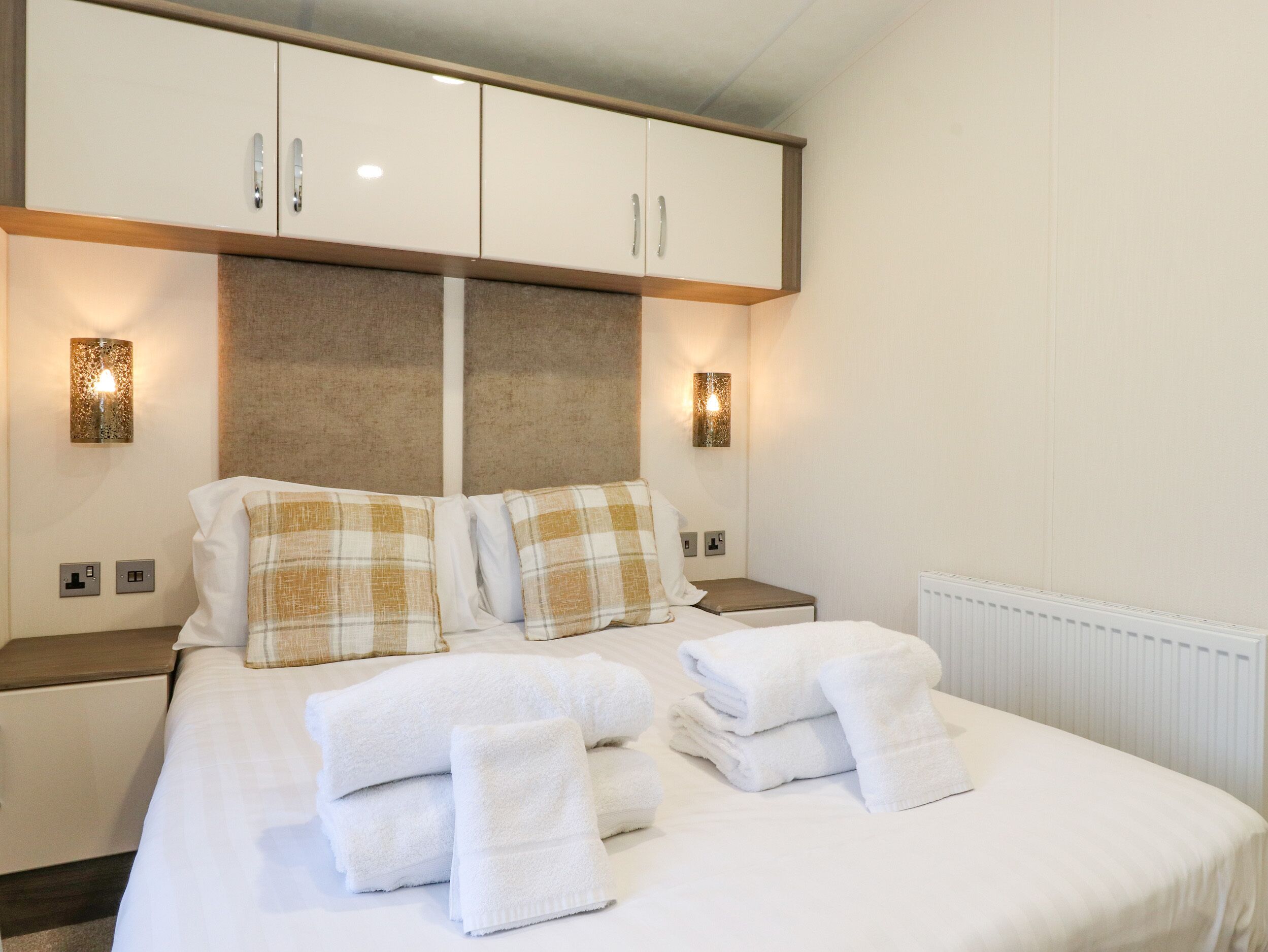 Un lit bien fait, avec du linge de lit blanc et des coussins à carreaux, une tête de lit en bois et deux tables de chevet avec des lampes.