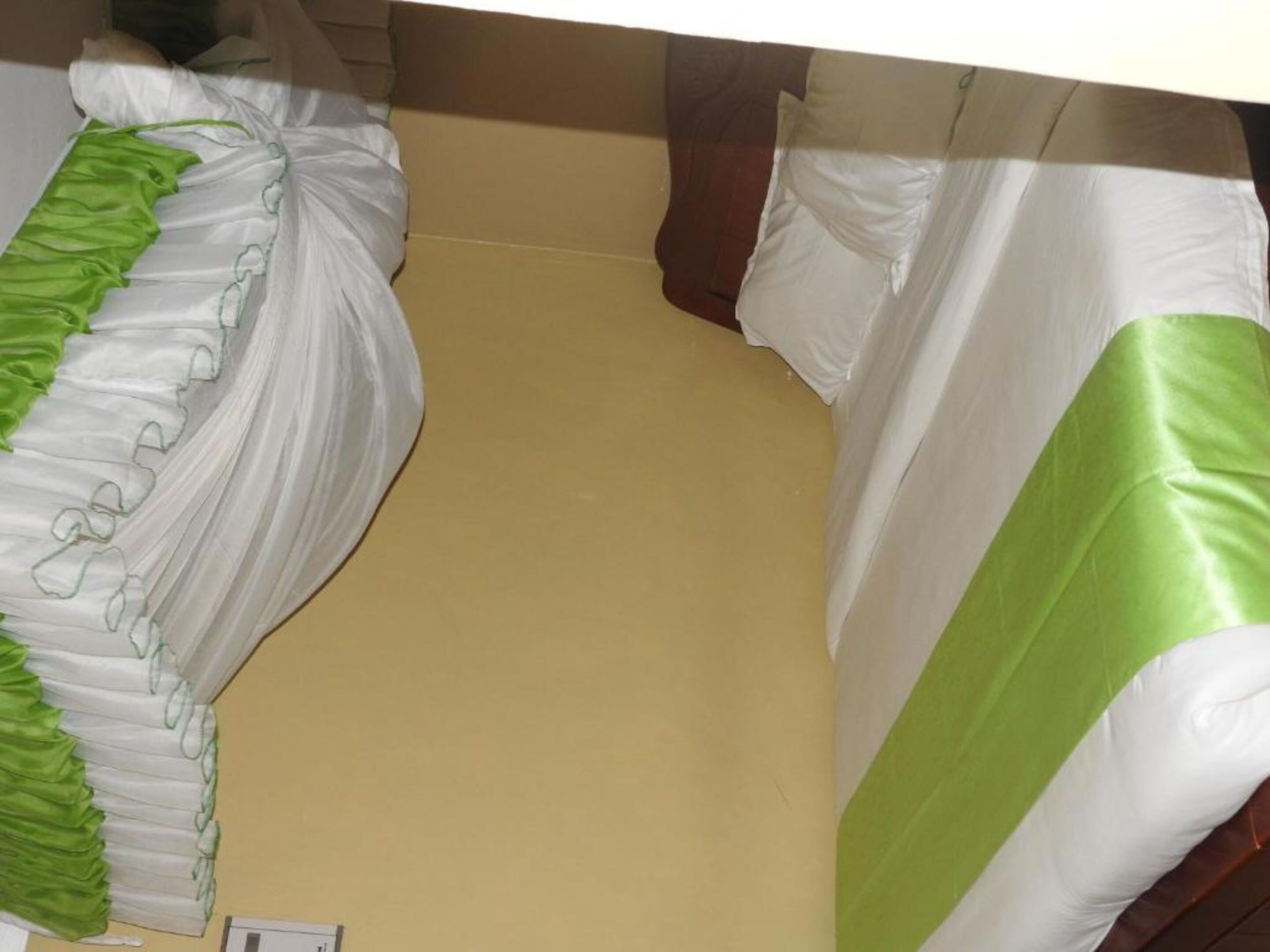 Une pièce avec un lit recouvert de draps blancs et verts, un mur beige et un petit objet blanc posé sur le sol.
