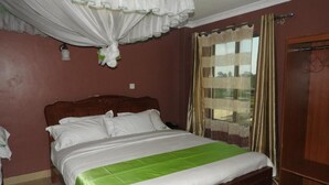 A bedroom with a wooden bed, white bedding, a green accent on the bedspread, a window with curtains, and a door. - Omega Gardens Hotel (Nanyuki)