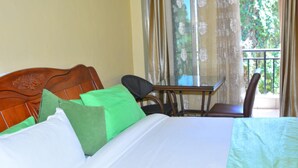 A hotel room with a wooden headboard, a bed with white and green bedding, a small table with chairs, and a view of greenery through a window with curtains. - Omega Gardens Hotel (Nanyuki)