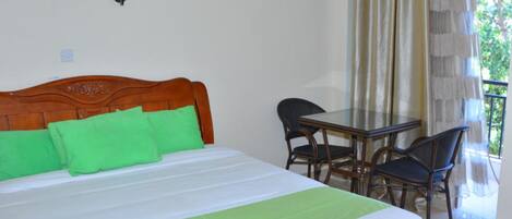 A bedroom with a wooden headboard, a bed with green pillows, a white bedspread, and a green blanket. There is a small table with chairs and a window with curtains.