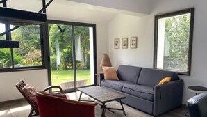 Ein modernes Wohnzimmer mit einem grauen Sofa, einem roten Sessel, einem Holztisch und Blick auf einen Garten durch große Fenster.