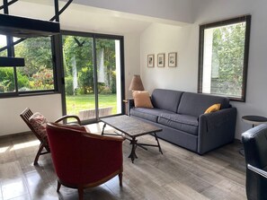 Un salon moderne avec un canapé gris, un fauteuil rouge, une table basse en bois et une vue sur un jardin grâce à de grandes fenêtres.