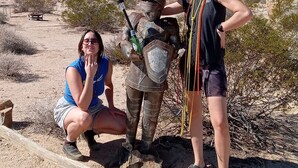 Two people in a desert setting, one in armor and the other in casual clothing, with a clear sky and sparse vegetation in the background.