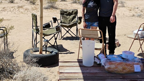 A desert camping setup with a wooden table, chairs, and an orange canopy.