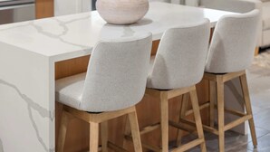 Une cuisine moderne avec un plan de travail en marbre blanc, une table en bois et trois tabourets de bar gris, recouverts de tissu.
