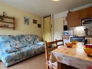Ein Wohnzimmer mit einer Couch, einem Esstisch mit Stühlen und einer Küchenzeile inklusive Mikrowelle, Herd und Schränken.