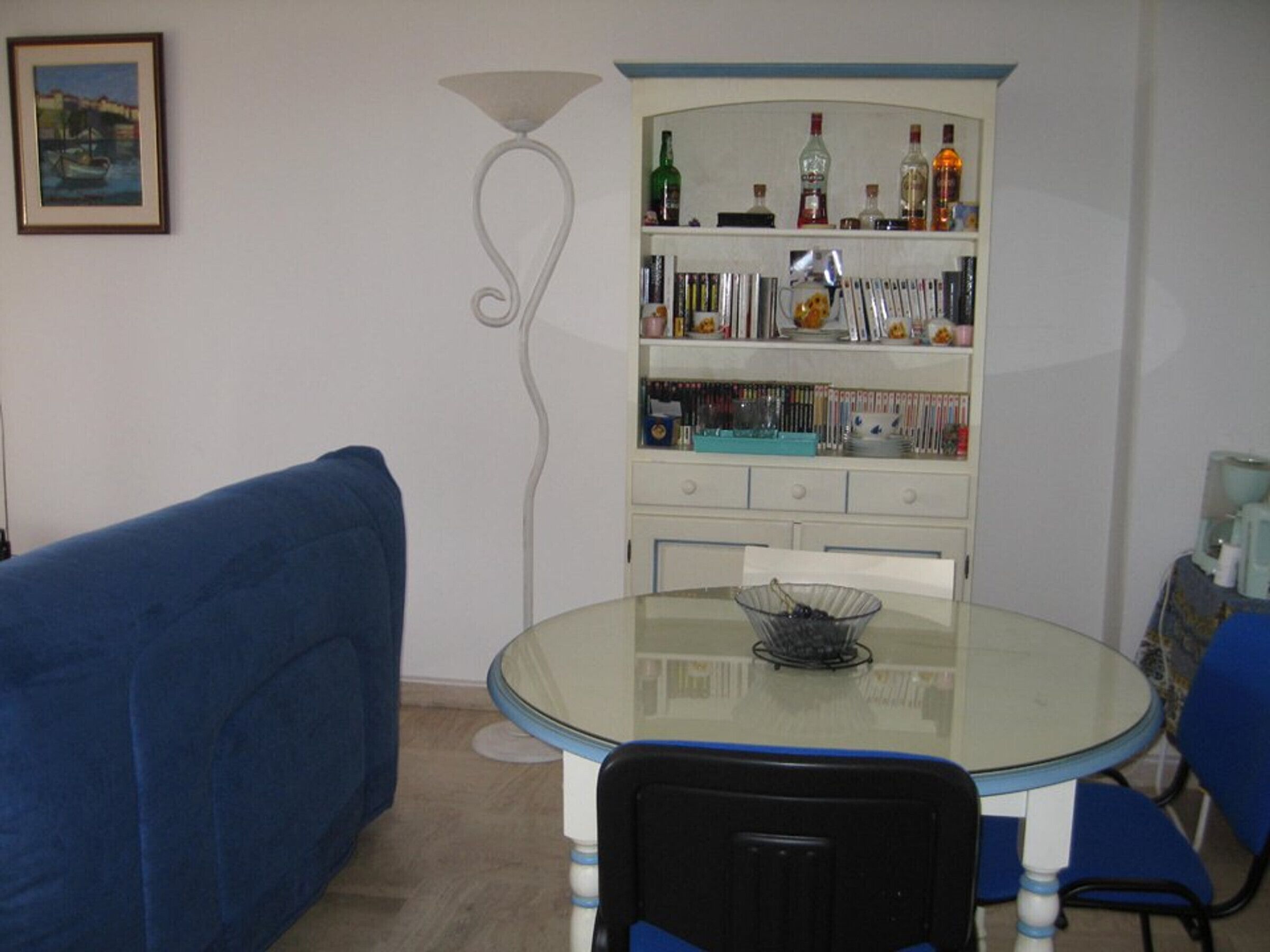 A dining area with a glass table, blue chairs, a white cabinet with bottles, and a framed picture on the wall.