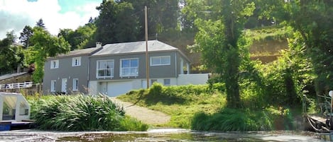A two-story house with a grey exterior and white trim, surrounded by lush greenery and a body of water.