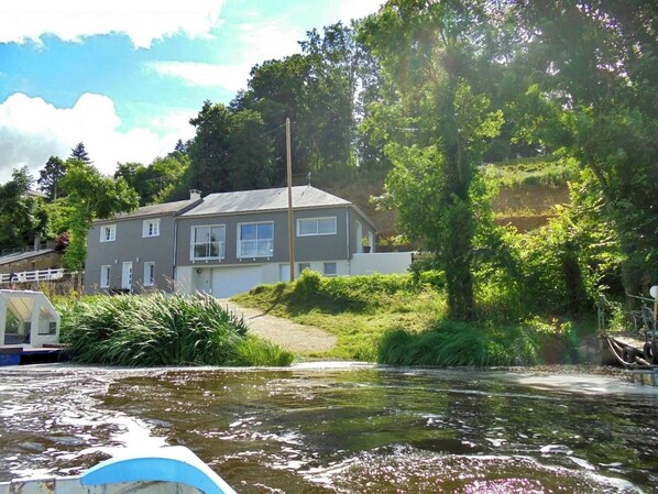 Une maison à deux étages, à l’extérieur gris et aux ornements blancs, entourée d’une végétation luxuriante et bordée d’un plan d’eau.