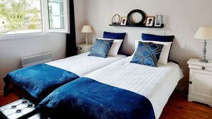 Een tweepersoonsbed met twee kussens, een nachtkastje met een lamp, een raam met gordijnen en een wandplank met decoratieve objecten.