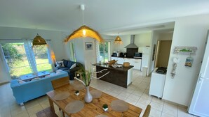 Eine moderne Küche mit einer zentralen Kochinsel, ein Essbereich mit einem Holztisch und gewebten Stühlen sowie ein Wohnbereich mit einem blauen Sofa und großen Fenstern.