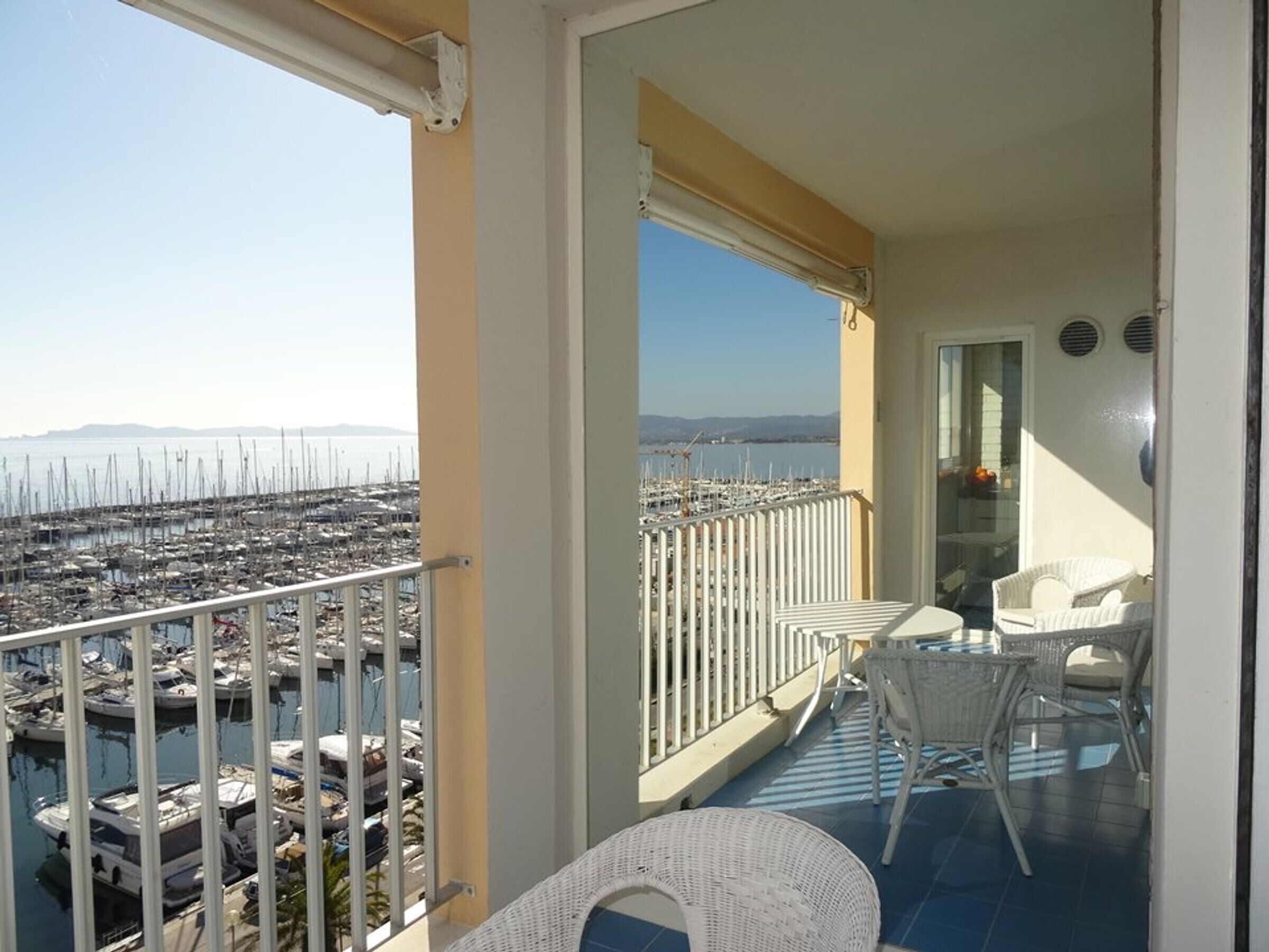 Ein Balkon mit Blick auf einen Yachthafen und Boote, weißen Korbmöbeln und einem gefliesten Boden.