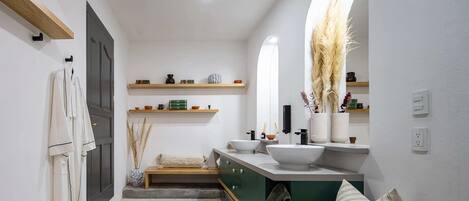 A modern bathroom with a large mirror, a white sink, and a vanity with a green cabinet. There are shelves above the sink holding decorative items and a towel rack on the left wall.