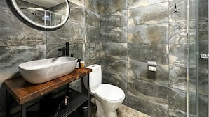 A bathroom with a marble wall, a round mirror, a wooden vanity, a white toilet, and a glass shower enclosure. - Beautiful 1-bed Apartment in Klaipeda (Klaipeda)