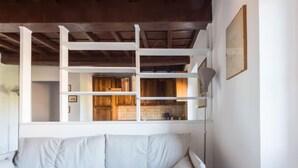 Ein weißes Sofa mit Kissen in einem Raum mit Holzboden und einer durch eine weiße Trennwand sichtbaren Küche.