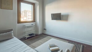 Ein Schlafzimmer mit einem Bett, einem kleinen weißen Regal, einem Fenster mit Aussicht und einem an der Wand befestigten Fernseher.