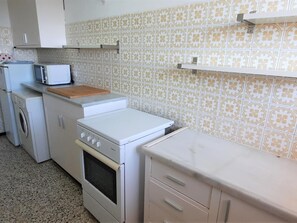 Cocina compacta con lavadora, microondas, estufa y una pequeña encimera con tabla de cortar.