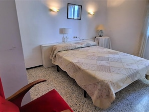A bedroom with a bed, a chair, a bedside table, a lamp, and a wall-mounted picture.