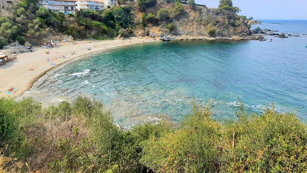Blick auf die Küste mit Strand, klarem Wasser und Gebäuden an einem Hang.