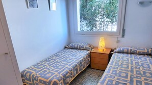 Ein kleines Schlafzimmer mit zwei Betten, einem Nachttisch mit Lampe, einem Fenster mit Blick ins Grüne und einer Tür.