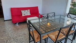 Ein Tisch mit Glasplatte, Stühle, ein rotes Sofa mit einem Blumenkissen, ein Bild an der Wand und eine Leuchte.