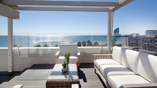 En balkon med en hvid sofa, et lille bord med en vase, og udsigt til havet og bybilledet.