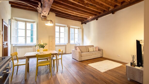 A spacious living room with a wooden ceiling, a sofa, a dining table with chairs, and a large window. - Dante´s Home Cozy in Center Town (Lucca)