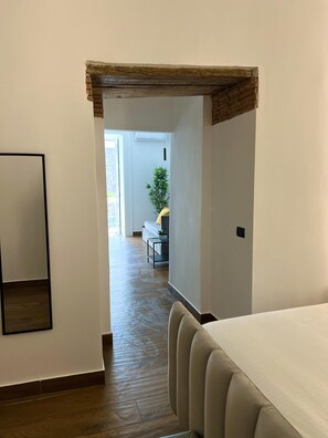 Et værelse med trægulv, en seng, et lille bord med en plante, og udsigt til et andet værelse med en sofa og en plante.