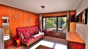 A cozy room with a wooden floor, a red patterned sofa, a small wooden table, and a window with a view of greenery.