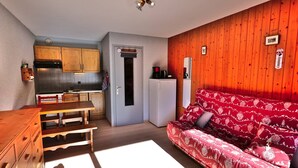 A compact living space with a kitchenette, a wooden dining table, a sofa with patterned cushions, and a door leading to another room.