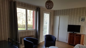 A living room with a TV on a wooden cabinet, two armchairs, a dining table with chairs, a white sofa, and a large window with curtains.