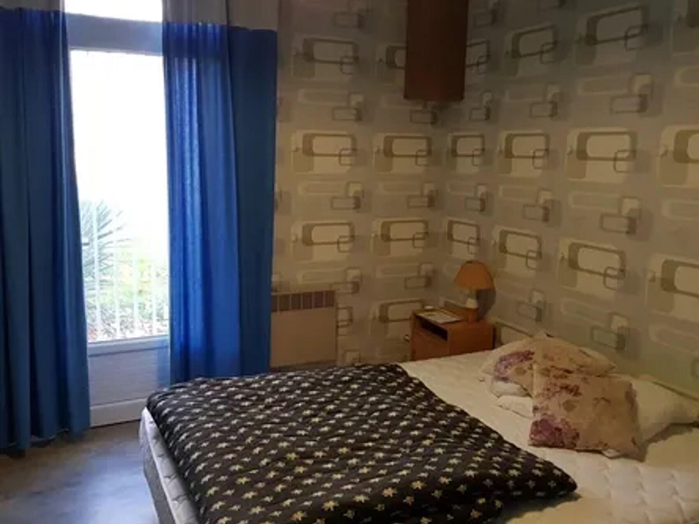 Un dormitorio con cama, mesita de noche, lámpara y ventana con cortinas azules.
