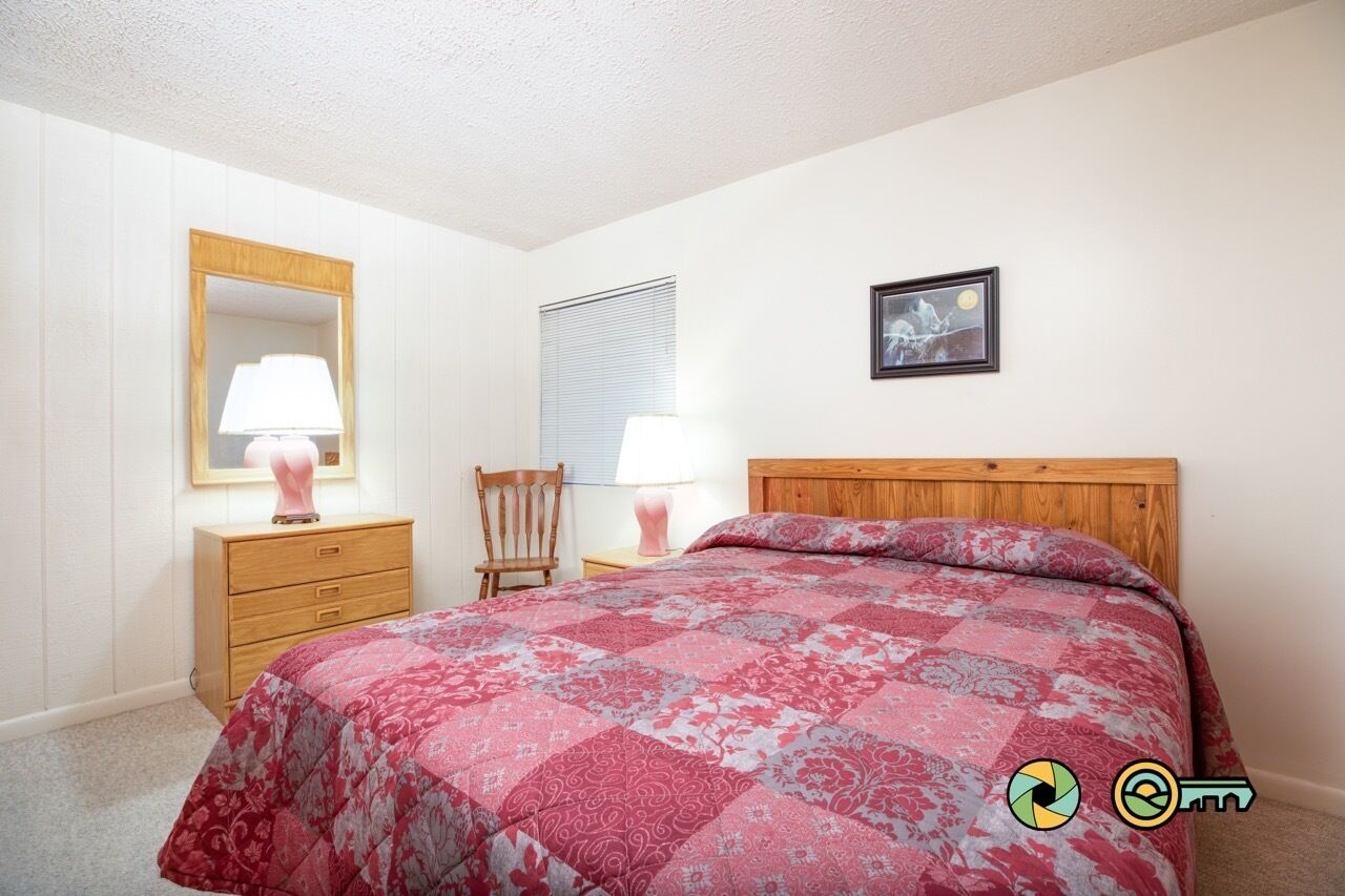 Ein Schlafzimmer mit einem Bett, einem Nachttisch aus Holz, einem Stuhl, einer Kommode, einem Spiegel, einem Fenster mit Jalousien und einem gerahmten Bild an der Wand.