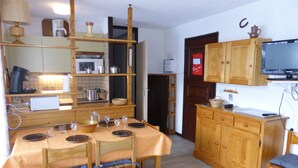 A compact kitchen with wooden cabinets, a dining table set for four, and a television mounted above the cabinet.