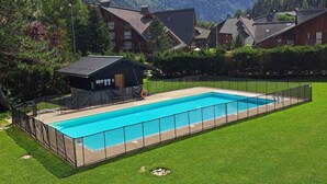 A rectangular swimming pool surrounded by a fence, with a small building and a grassy area in the foreground, and mountains in the background.