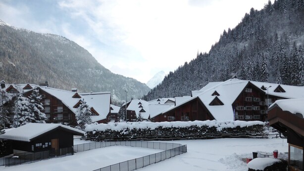 A snow-covered lodge with multiple chalet-style buildings, a fenced courtyard, and a mountainous backdrop.