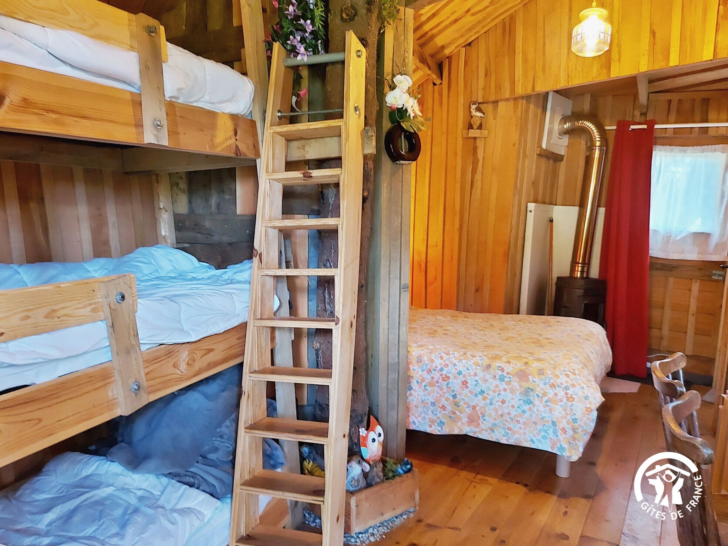 Une pièce rustique en bois, avec des lits superposés, une échelle, une petite étagère ornée d’objets décoratifs et une fenêtre avec des rideaux.