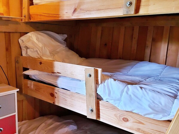 Una cama litera de madera con una cama individual en la parte superior y una inferior. A la izquierda de la cama hay un pequeño aparador de madera.