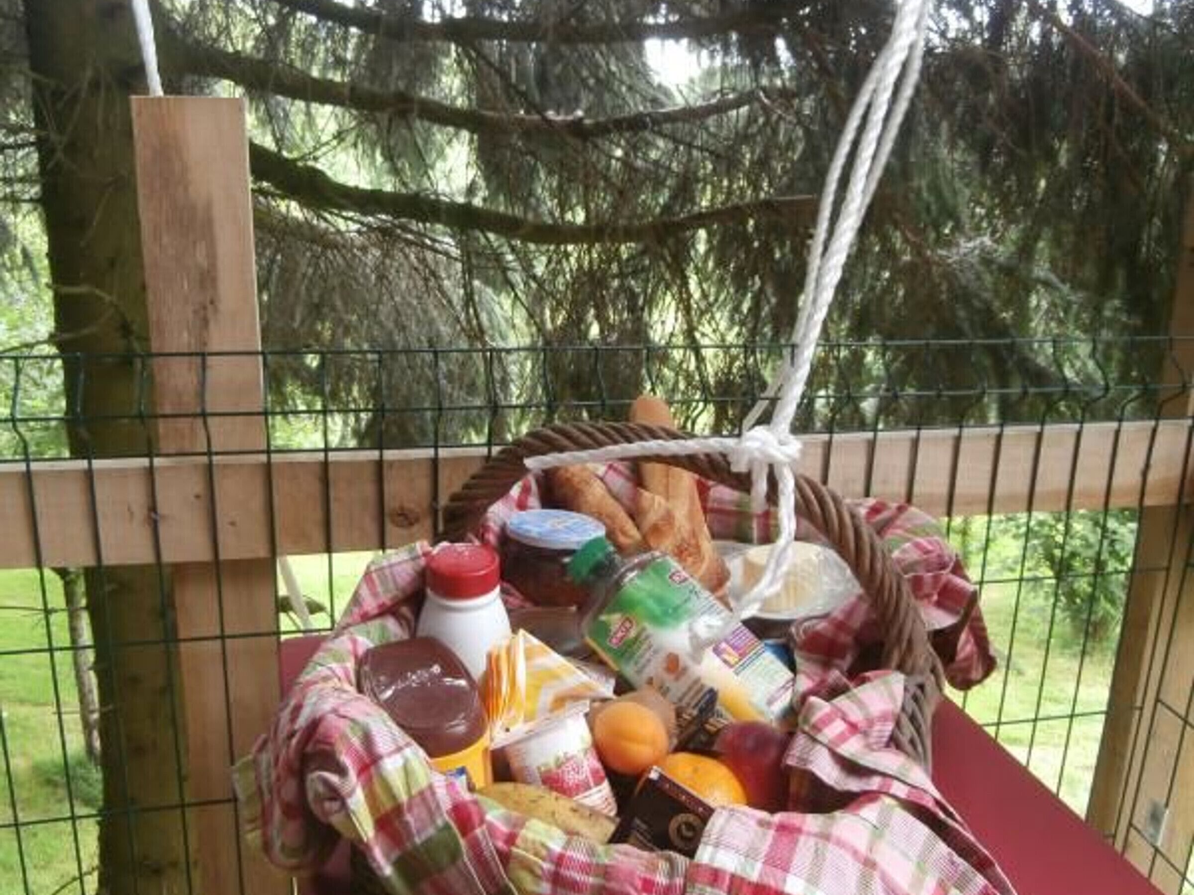 Una cesta de picnic llena de comida, como pan, frutas y salsas, colgada de una valla de madera con un paisaje natural de fondo.