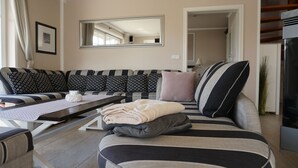 A modern living room with a sofa, a coffee table, and a dining table. There is a mirror on the wall and a door leading to another room.