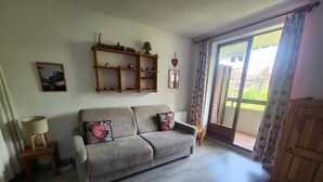 A living room with a grey sofa, wooden shelves, a small table with a lamp, and a view through a sliding glass door. - Cozy studio in village center, pets allowed, private parking (Samoëns)