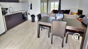 A modern kitchen with dark cabinets, a built-in oven, and a sink. There is a dining area with a table and chairs, and a view of a garden through a window. - Charming Renovated Stone House on a Farm with Modern Amenities, Near Scenic Trails (Champéon)