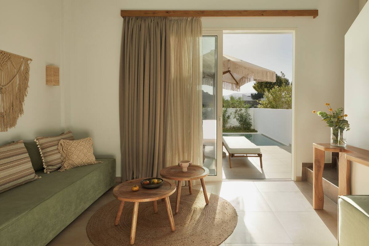 Una sala moderna con un sofá verde, dos mesas de madera, un tapete tejido y un amplio ventanal corredero que da a un área exterior con piscina.