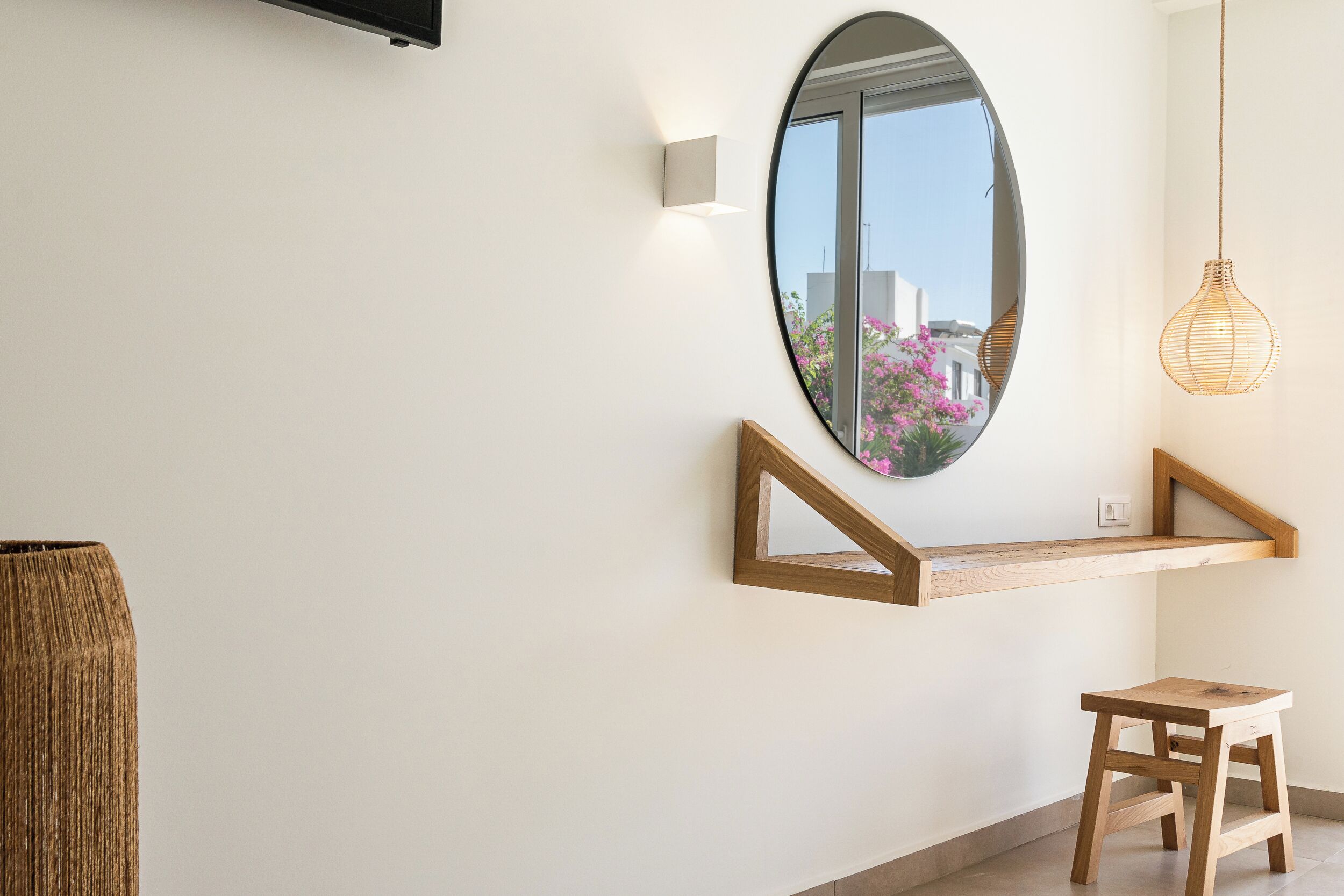 A modern interior with a wooden bench, a small stool, a wall-mounted shelf, and a round mirror reflecting a pink flower outside.