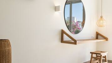 A modern interior with a wooden bench, a small stool, a wall-mounted shelf, and a round mirror reflecting a pink flower outside.