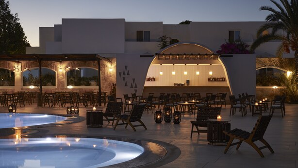 Un área de comedor al aire libre con mesas y sillas, una piscina y un bar con techo semicircular.