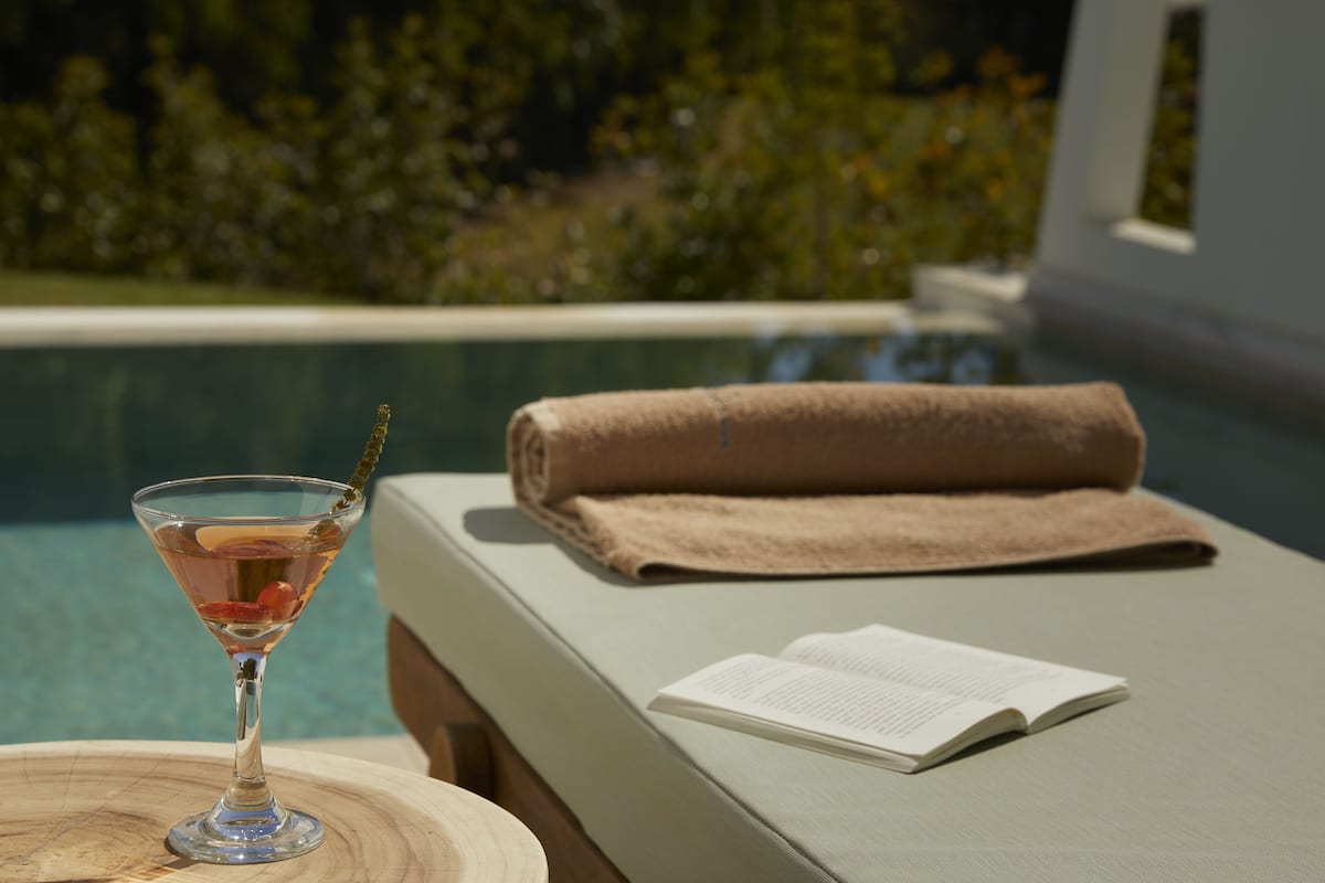 Una vista despejada de un área de descanso junto a la piscina, con una bebida rosada, un libro y una toalla sobre un banco acolchado.